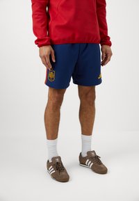 Mornarske atletske kratke hlače s žutim Adidas logotipom, bočnim trakama i grbom Španije. Uparene s smešnim cipelama i bijelim čarapama.