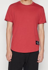 Camiseta de algodón roja con escote redondo y mangas cortas enrolladas. Presenta un parche del logo de Calvin Klein en negro en la parte inferior izquierda.