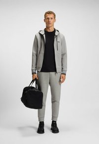 Felpa grigia con zip e dettagli neri, abbinata a una t-shirt nera a girocollo e pantaloni della tuta grigi coordinati. Borsa a tracolla nera e sneakers.