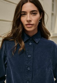 Granatowa koszula z korduroyu z zapięciem na guziki, kołnierzem rozłożonym i teksturowanym materiałem. Posiada sześć dopasowanych niebieskich guzików wzdłuż listwy guzika.