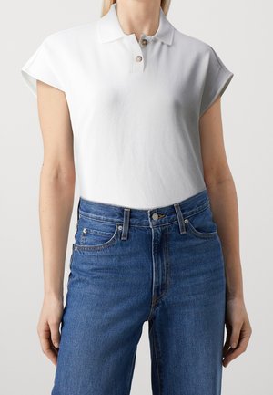 Persoon draagt een wit poloshirt met korte mouwen en twee knopen, en een hoge taille blauwe spijkerbroek tegen een effen achtergrond.