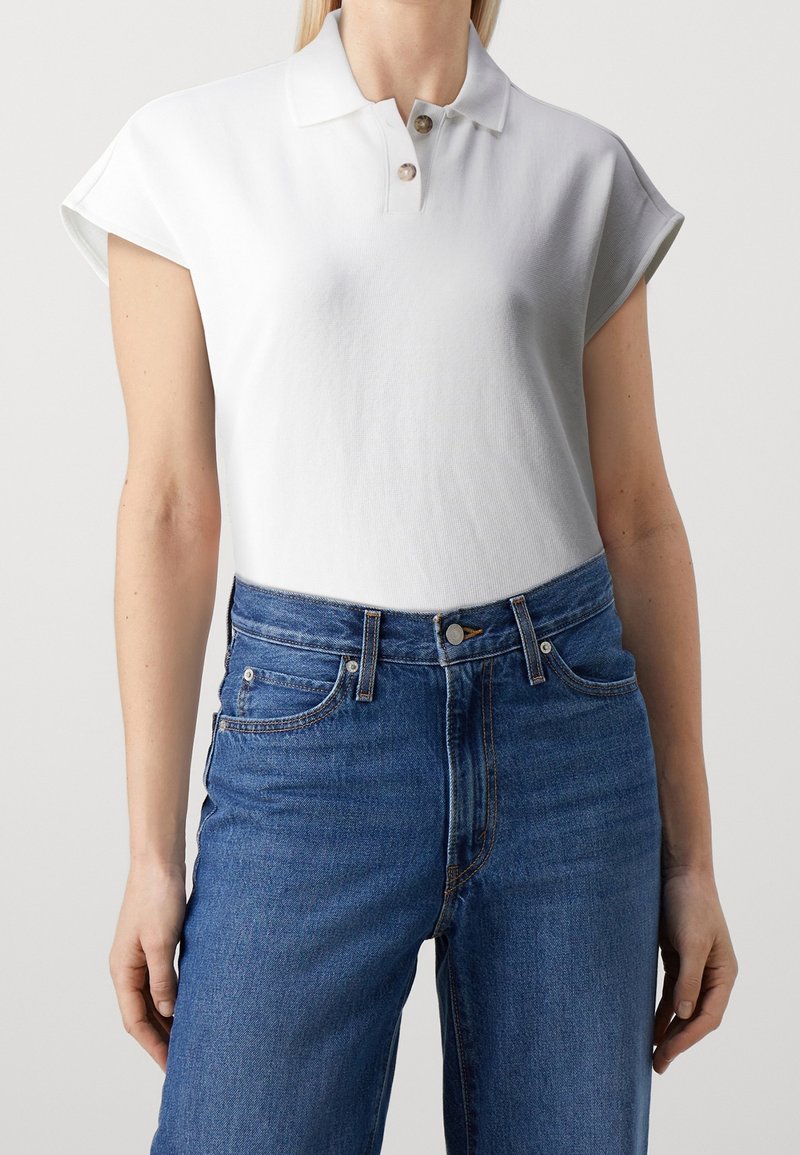 Personne portant un polo blanc à manches courtes avec deux boutons et un jean bleu taille haute sur un fond uni.