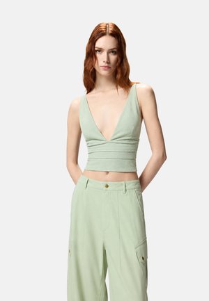 Giovane donna con capelli rossi che indossa un crop top verde chiaro senza maniche con scollo a V profondo e pantaloni larghi a vita alta coordinati, in piedi davanti a uno sfondo bianco.