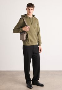 Olijfgroene hoodie met een voorzak, gedragen met zwarte broek en zwarte schoenen. Het model houdt een kleine bruine schoudertas vast.