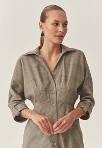 Vestito a quadri marrone con bottoni, colletto, maniche lunghe arrotolate ai polsini e bottoni di legno. Tessuto liscio con una vestibilità strutturata.