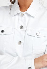 White denim jacket featuring a collar, silver buttons, and two chest pockets with flaps. Smooth texture and classic design.