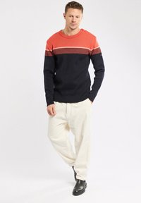 Homme portant un pull rayé rouge et noir, un pantalon crème et des bottes noires, se tenant les mains dans les poches contre un fond clair.