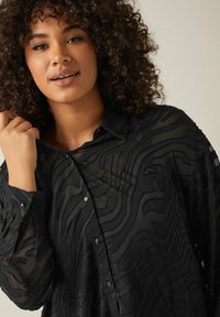 Black shirt made of sheer fabric featuring a textured wave pattern. It has a collared neck and button-down front with slight translucent details.