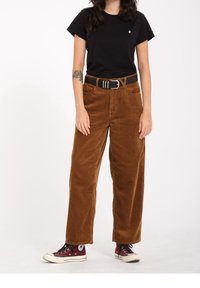 Kvinde iført en sort t-shirt, brune fløjl bukser med et sort bælte og røde høj sneakers, stående foran en hvid baggrund.
