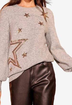 Grijze trui met gouden paillettensterren gecombineerd met een hoge taille donkerbruine leren broek bij een staand persoon.