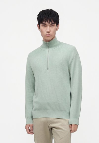 Jeune homme portant un pull vert clair à col montant zippé et un pantalon beige, debout devant un fond blanc uni.