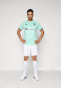 Mintgrünes Fußballtrikot mit schwarzen Akzenten und Logo; weiße Shorts; weiße Socken; schwarz-weiße Schuhe; glatte Textur, athletic Fit.
