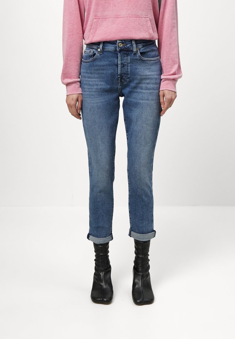 Vaqueros azules ajustados con puños enrollados, hechos de mezclilla. Combinados con botas de tobillo negras y holgadas y una sudadera rosa de manga larga.
