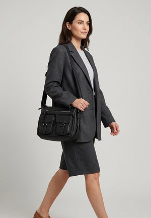 Kobieta w szarym garniturze biznesowym idzie, niosąc czarną skórzaną torebkę na ramię z dwoma przednimi kieszeniami, na jednolitym jasnym tle.