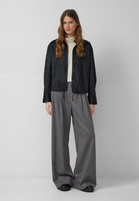 Kobieta o długich falowanych włosach, ubrana w czarną zapinaną na guziki kurtkę, kremowy golf, szare spodnie w szerokich nogawkach w prążki i czarne buty, stoi na tle szarej ściany.