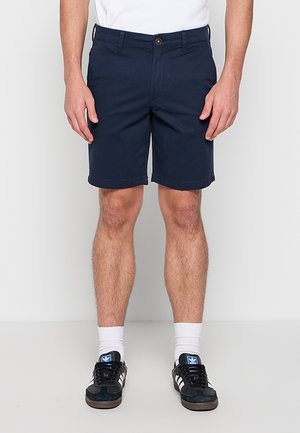 Man draagt marineblauwe korte broek, witte sokken en zwarte Adidas sneakers met witte strepen, staand tegen een effen lichte achtergrond.