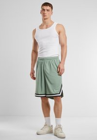 Lysgrønne basketballshorts med sorte accenter, parret med en hvid tanktop og beige sneakers, vist på en mandlig model.