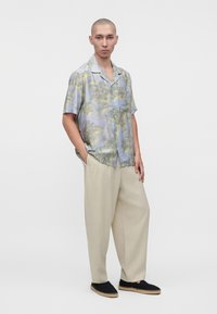 Hombre de pie con las manos en los bolsillos, vestido con una camisa de flores pastel de manga corta, pantalones beige de pierna ancha y alpargatas negras.
