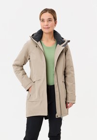 Beige waterdichte jas met een zwarte capuchon, voorzien van een rits aan de voorkant, twee voorzakken en een losse pasvorm, gedragen over een groene shirt.