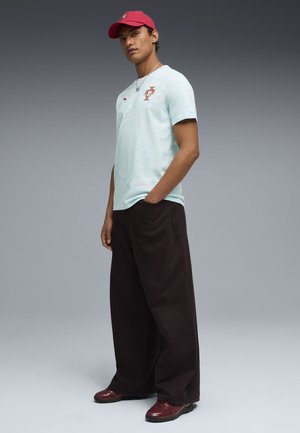 Jonge man die een lichtblauw voetbalshirt van Portugal draagt, een rode pet, zwarte wijde broek en rode sneakers, staand met één hand in zijn zak.