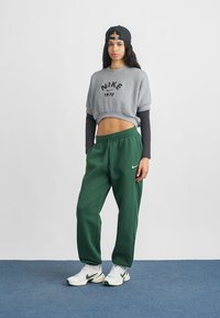 Γκρίζα κοντή φόρμα με μαύρο λογότυπο "Nike 1972", συνδυασμένη με πράσινα παντελόνια φόρμας και λευκά αθλητικά παπούτσια με πράσινες λεπτομέρειες. Καπέλο επίπεδο φορεμένο στο πλάι.