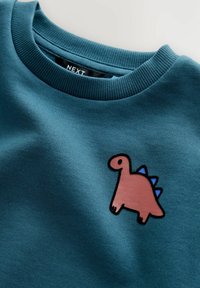 Felpa in cotone teal con collo rotondo, caratterizzata da un'applicazione di un dinosauro rosa con spuntoni blu, messa in risalto da una linea nera sul petto.