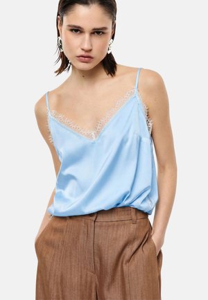 Donna che indossa una canotta di raso azzurro chiaro con bordatura in pizzo e pantaloni marroni a vita alta, mani nelle tasche, espressione neutra, capelli corti.