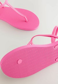Vaaleanpunaiset kumiset flip-flopit, joissa on punottu remmi ja teksturoidut pohjat. Pohjassa on uritettu kuvio, joka parantaa pitoa, sekä pyöreä koristetieto.