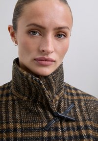 Abrigo de lana a cuadros marrón y negro con un cuello alto, que cuenta con un cierre de toggle negro. Textura suave con una apariencia cálida.