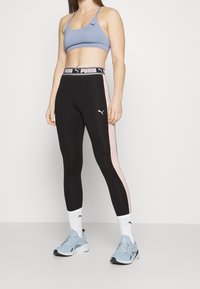 Zwarte legging met een roze zijpaneel, gecombineerd met een lichtblauwe sportbeha met een zwart logo. Witte enkelsokken en lichtblauwe sneakers maken de look af.