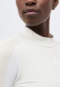 Κοντινό πλάνο ενός ατόμου που φοράει λευκό αθλητικό τοπ με ψηλό γιακά, διάτρητο μανίκι και ανάγλυφο λογότυπο στο στήθος.