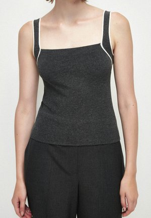 Femme portant un haut sans manches gris foncé ajusté avec une bordure blanche sur les bretelles, associé à un pantalon ample gris foncé, sur un fond uni.