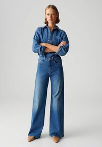 Chemise en denim avec patte de boutonnage, manches longues et poche poitrine, associée à un jean bleu délavé à jambes larges. Des chaussures de couleur neutre complètent la tenue.