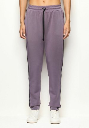 Persona vestida con pantalones de chándal morados con franjas laterales negras y zapatillas blancas, de pie contra un fondo claro y liso.