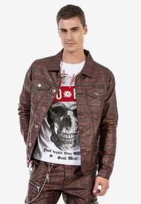 Rot-schwarz kariertes Jeansjackett mit Fronttaschen und Metallnieten. Über einem weißen T-Shirt mit grafischem Aufdruck, das ein Skull-Motiv und Text zeigt, getragen.