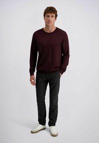 Mann trägt einen dunkelburgunderfarbenen Pullover mit langen Ärmeln, dunkelgraue Hosen und weiße Sneakers, steht mit einer Hand in der Tasche vor einem schlichten Hintergrund.