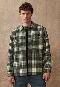 Groen-wit geruit flanellen overhemd met knoopsluiting, voorzien van een kraag en borstzakken, gecombineerd met donkere jeans.