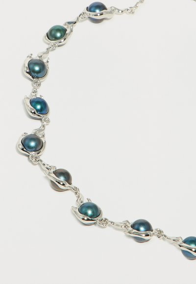 Ασημένιο κολιέ με συνδεδεμένες ιριδίζουσες σκούρες μπλε πέρλες σε οργανικές σχηματισμένες θήκες σε λευκό φόντο.