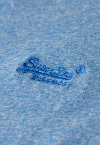 Blauwe stof met geborduurd "SuperDry"-logo in donkerblauwe draad, gecentreerd in het beeld.