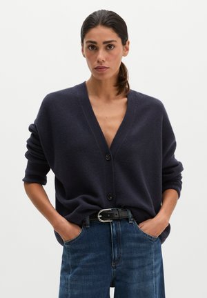 Junge Frau mit dunklem Pferdeschwanz, die eine marineblaue Strickjacke mit hochgekrempelten Ärmeln und blaue Jeans trägt, Hände in den Taschen, neutraler Hintergrund.