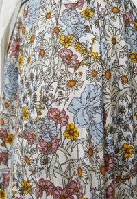 Tissu floral avec marguerite détaillée, fleurs bleues, roses et jaunes sur fond clair, montrant la texture et le design du motif.