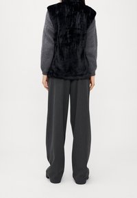 Gilet in ecopelliccia nero con collo alto e maniche grigie lavorate a maglia, abbinato a pantaloni larghi neri a righe sottili.