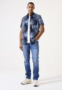 Camisa de corto con patrón en azul marino y celeste con hojas de palmera, combinada con jeans celestes y zapatillas beige. El modelo posa de pie.