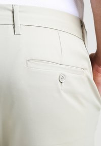 Pantaloni beige chiaro con una tasca posteriore con chiusura a bottone. Il tessuto è liscio con una leggera lucentezza e un design sartoriale.
