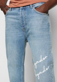Ljust blå denimjeans med avslappnad passform, som har två framfickor, blekt textur och vit tryckt text på vänster lår.