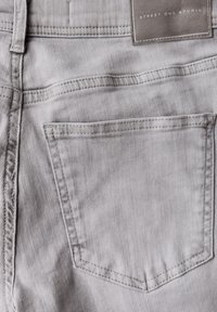 Světle šedé džíny z denimu s náprsní kapsou, jemnou texturou a přišitým logem na pásku. Jednoduchý design, bez ozdob.