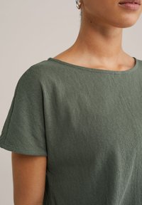 Tumši zaļš, īsām piedurknēm tops, izgatavots no mīksta, viegla auduma. Tam ir apaļa kakla izgriezums un brīvā piegriezuma forma ar minimālām šuvju detaļām.