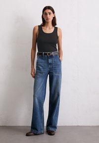 Débardeur gris à encolure dégagée, associé à un jean en denim bleu à jambes larges. Une ceinture en cuir marron et des bottines complètent la tenue.