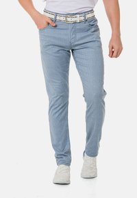 Gestreifte blaue Hose mit einer taillierten Passform, aus leichtem Stoff gefertigt. Verfügt über einen weißen Gürtel mit schwarzen Nieten, kombiniert mit hellen Schuhen.