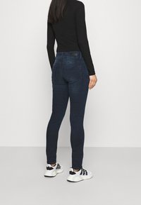 Mörkblå tajta jeans med medelhög midja, två bakfickor och subtil textur, tillsammans med en svart långärmad tröja och vita sneakers.